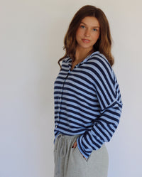 A woman wearing a navy and light blue horizontally striped long-sleeve button-up cardigan and light gray sweatpants, standing against a plain off-white wall.