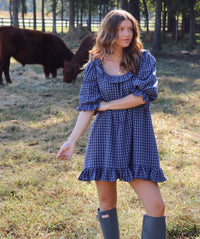 A woman wearing a blue gingham puff-sleeve dress with a ruffled neckline and hem, standing in a grassy field with cows in the background. She is wearing rain boots.