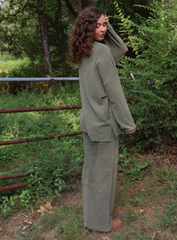 A woman wearing a matching olive green striped lounge set stands outdoors in a wooded area with greenery and a rustic fence behind her.