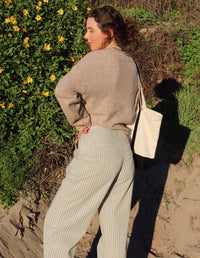 Woman wearing a beige sweater top and striped pants standing in front of greenery with yellow flowers.