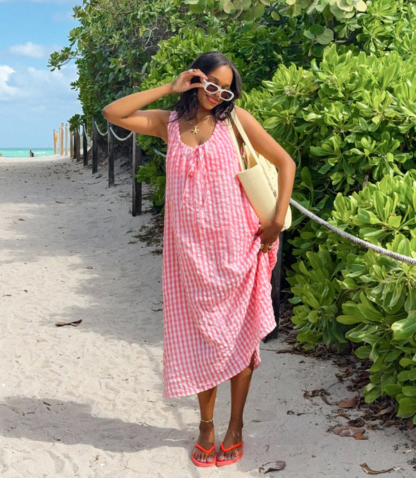 Woman in a pink checkered dress standing on a sandy path with greenery and ocean in the background