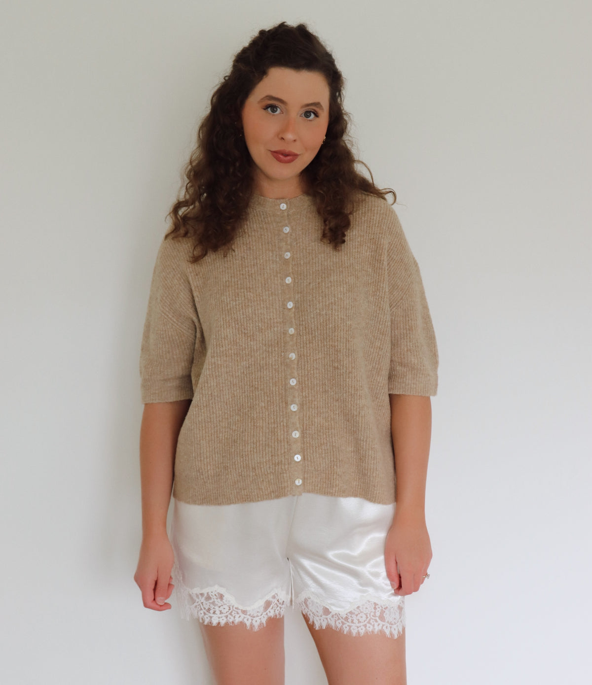 A woman modeling a beige short-sleeve button-front cardigan sweater paired with white satin shorts featuring a lace hem, set against a plain white wall.
