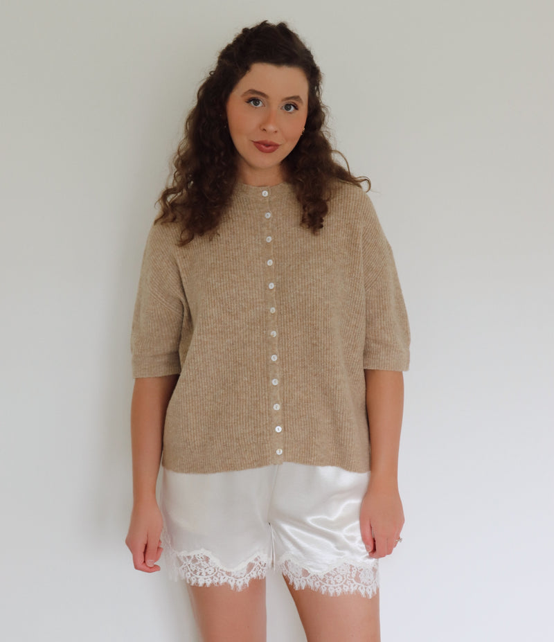 A woman modeling a beige short-sleeve button-front cardigan sweater paired with white satin shorts featuring a lace hem, set against a plain white wall.