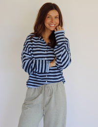 A woman wearing a navy and light blue horizontally striped long-sleeve button-up cardigan and light gray sweatpants, standing against a plain off-white wall.