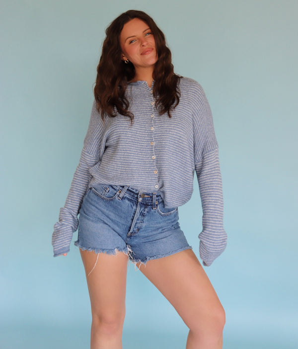 Woman wearing a striped long-sleeve cardigan and denim shorts against a light blue background