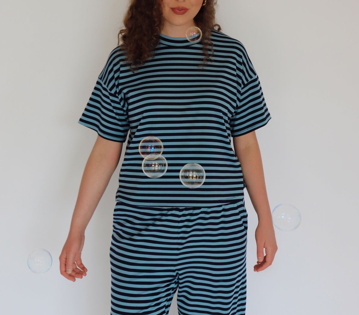 A woman wearing a matching two-piece loungewear set with dark teal and navy blue horizontal stripes, standing against a plain white wall with floating bubbles.