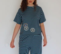 A woman wearing a matching two-piece loungewear set with dark teal and navy blue horizontal stripes, standing against a plain white wall with floating bubbles.