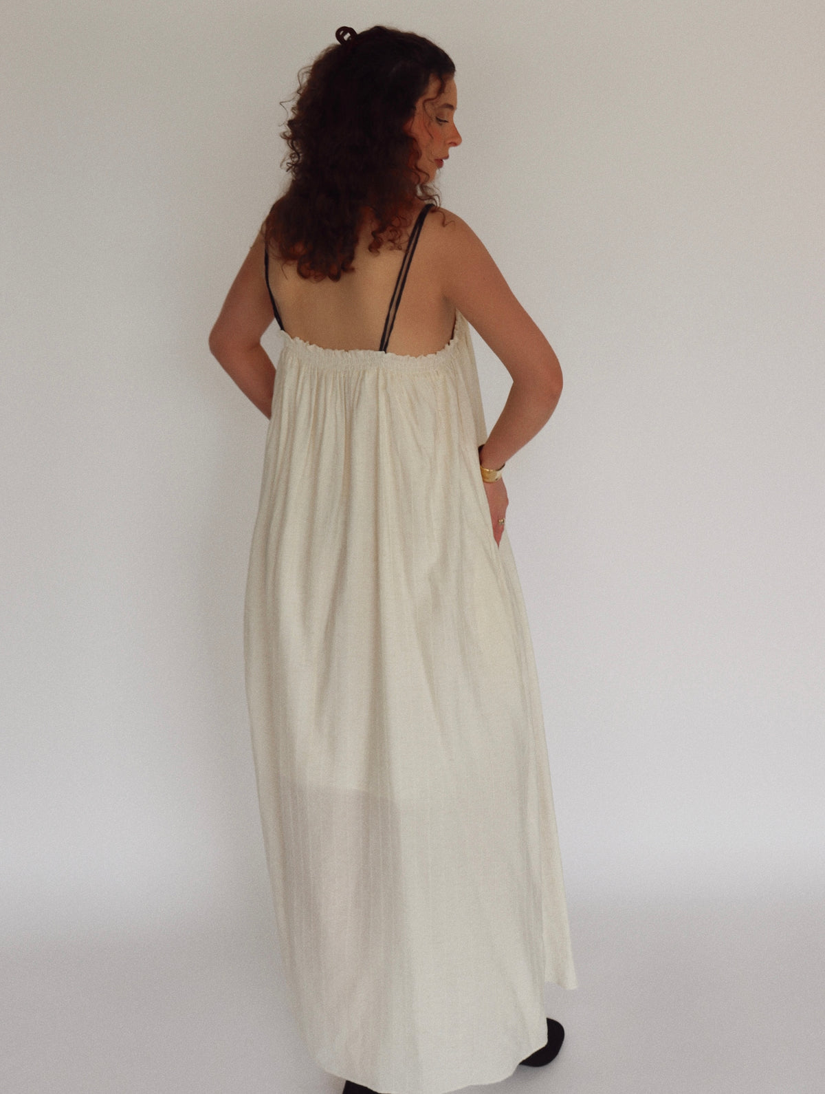 Woman wearing a long, light-colored dress with black straps against a plain background