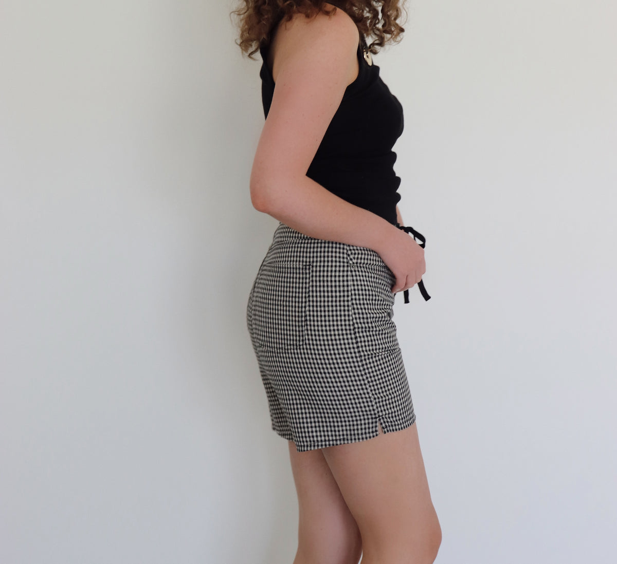 A woman with curly brown hair wearing a black tank top and black-and-cream gingham shorts, standing against a plain white wall.