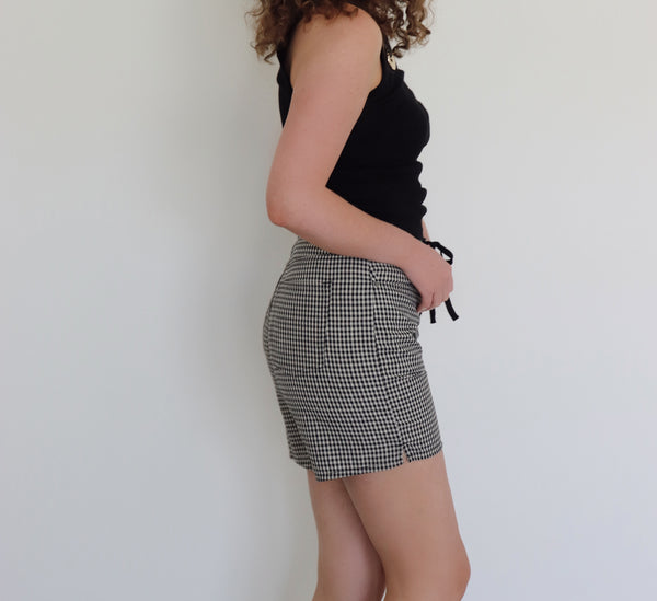 A woman with curly brown hair wearing a black tank top and black-and-cream gingham shorts, standing against a plain white wall.