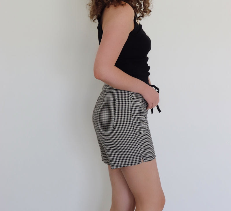 A woman with curly brown hair wearing a black tank top and black-and-cream gingham shorts, standing against a plain white wall.