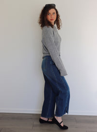 Woman wearing a gray sweater top and blue jeans standing against a white wall.