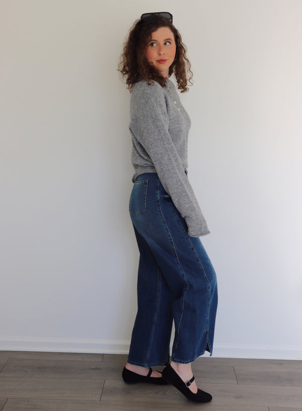 Woman wearing a gray sweater top and blue jeans standing against a white wall.