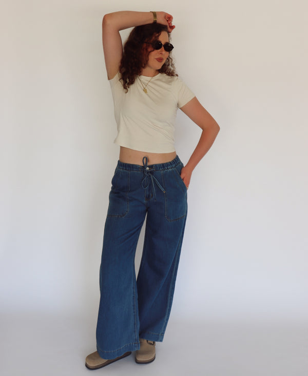 A woman modeling a cream short-sleeve crop top and wide-leg blue jeans against a plain white studio background.