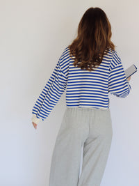 A woman shown from behind wearing a blue and cream horizontally striped long-sleeve crop top and light gray sweatpants, holding a book against a plain white wall.