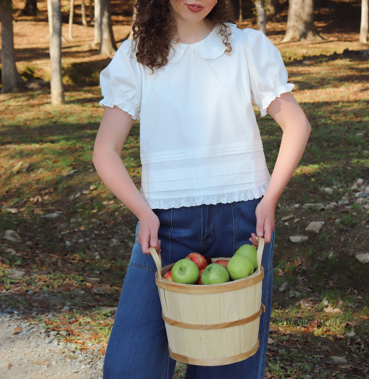 A woman with curly brown hair stands outdoors in a wooded area holding a wooden basket filled with green and red apples. She is wearing a white short-sleeve blouse with a ruffled collar and dark blue pants.