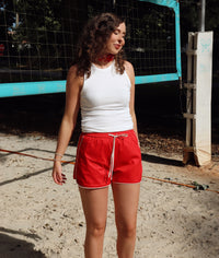 Woman in white tank top and red shorts standing on a sandy surface with a volleyball net in the background.