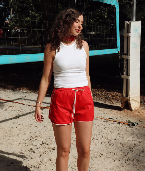 Woman in white tank top and red shorts standing on a sandy surface with a volleyball net in the background.