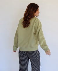 A woman with long brown hair is shown from behind wearing a light sage green crewneck sweatshirt and navy and white striped pants, standing against a plain white wall.