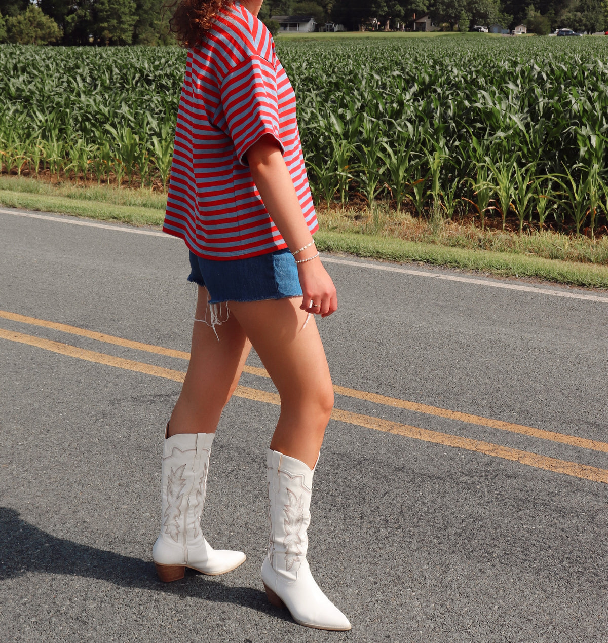 Person wearing a red and blue striped shirt, blue shorts, and white cowboy boots standing on a road with a cornfield in the background.