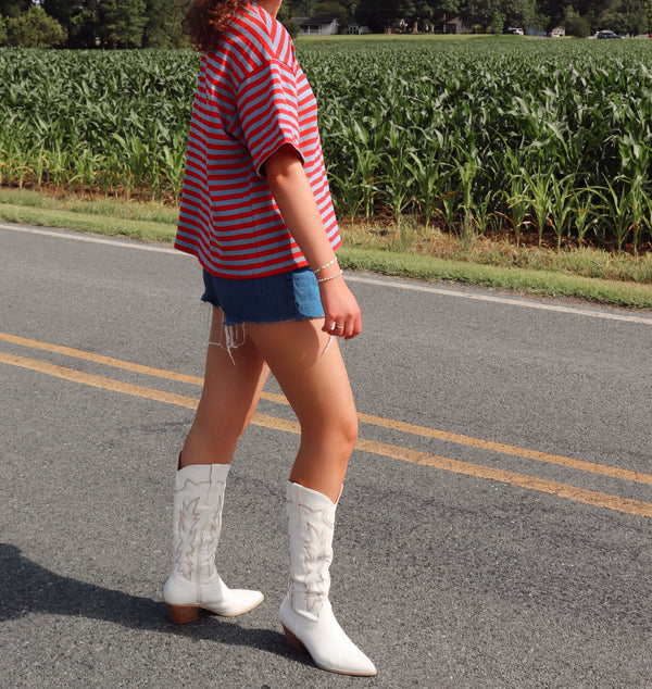 Person wearing a red and blue striped shirt, blue shorts, and white cowboy boots standing on a road with a cornfield in the background.