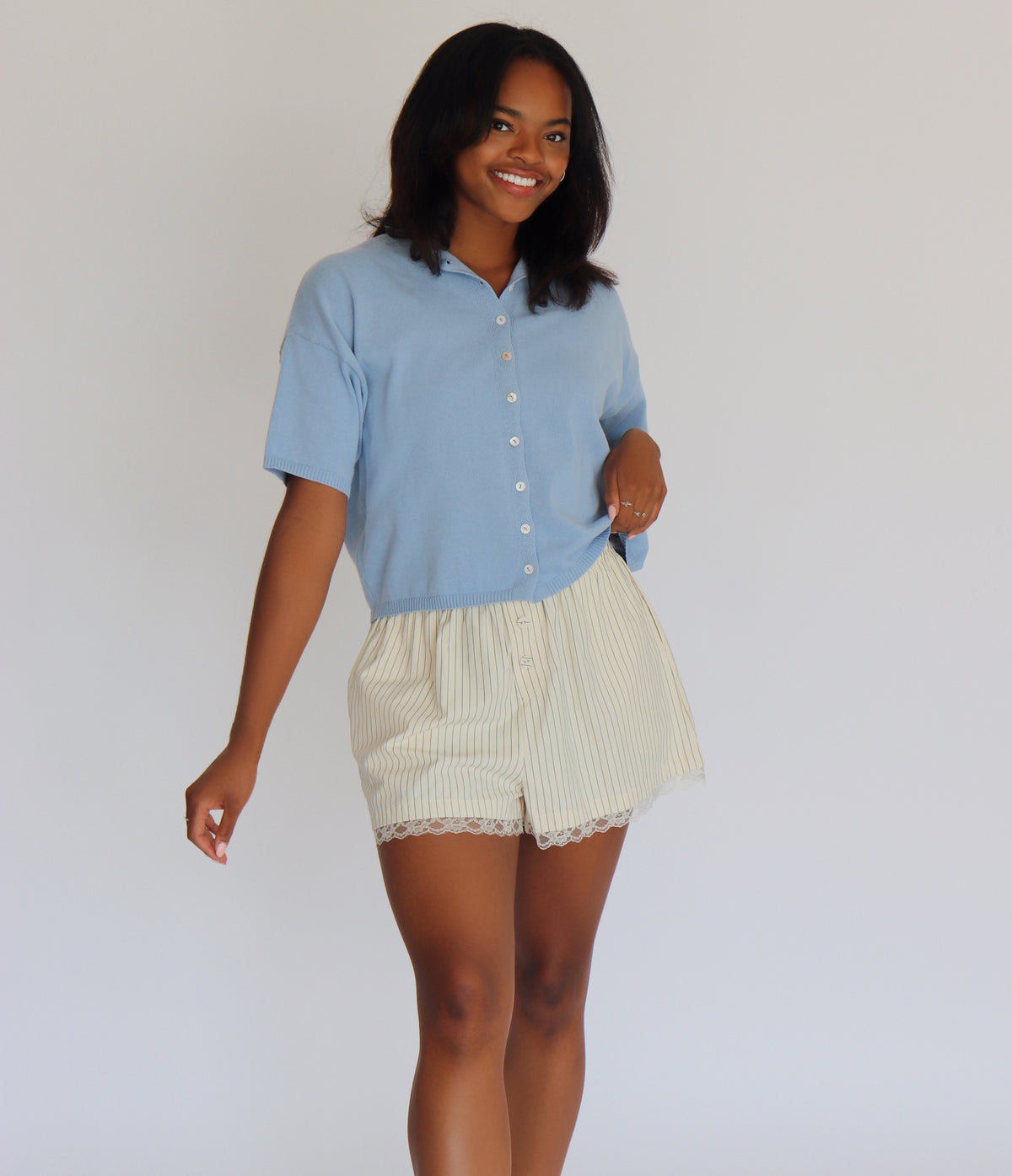 Woman wearing a blue short sleeve cardigan and cream shorts against a white background