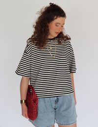 Woman wearing a black and white striped shirt with denim shorts and holding a red bag against a plain background