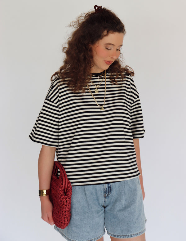 Woman wearing a black and white striped shirt with denim shorts and holding a red bag against a plain background