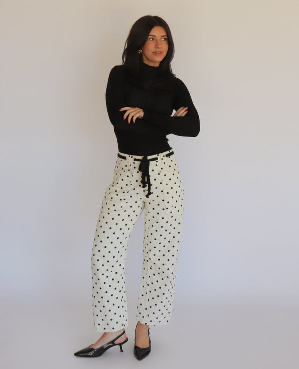 A woman in a black turtleneck and white barrel jeans with black polka dots on them, in front of a white background