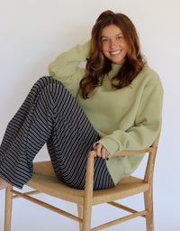 A smiling woman with long brown hair sits on a light wood chair with a woven seat. She is wearing an oversized sage green mock-neck sweatshirt and navy-and-white vertically striped wide-leg pants against a plain white wall.