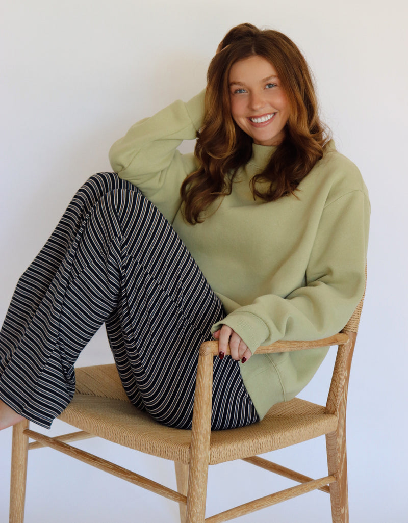 A smiling woman with long brown hair sits on a light wood chair with a woven seat. She is wearing an oversized sage green mock-neck sweatshirt and navy-and-white vertically striped wide-leg pants against a plain white wall.