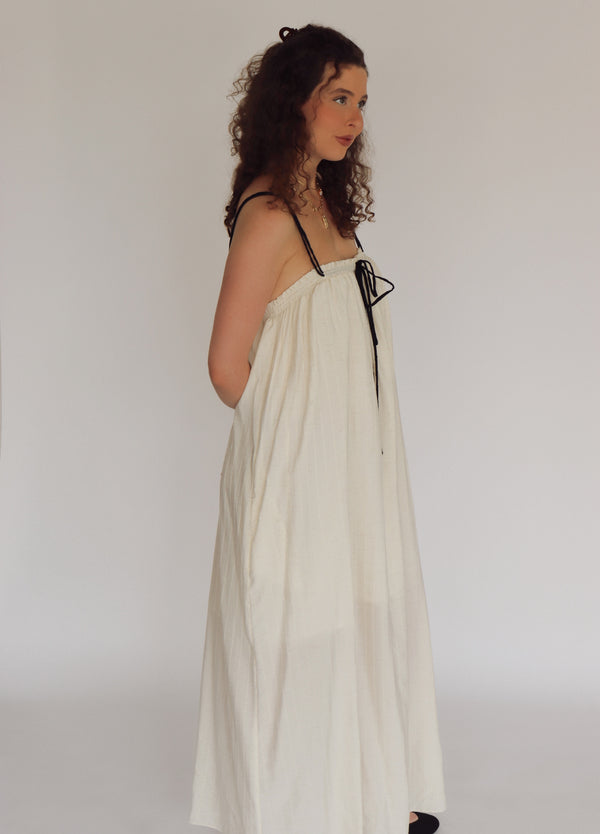 Woman wearing a long, light-colored dress with black straps against a plain background