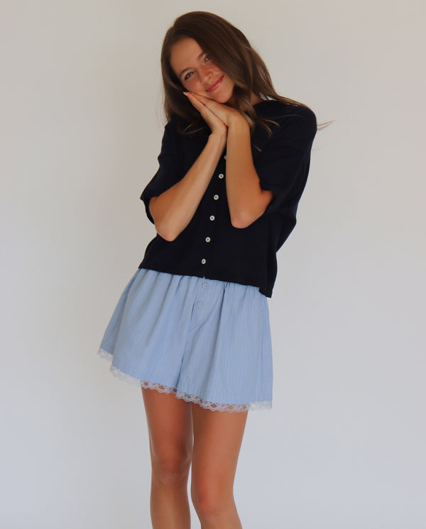 Woman wearing a navy short sleeve cardigan and light blue shorts against a white background