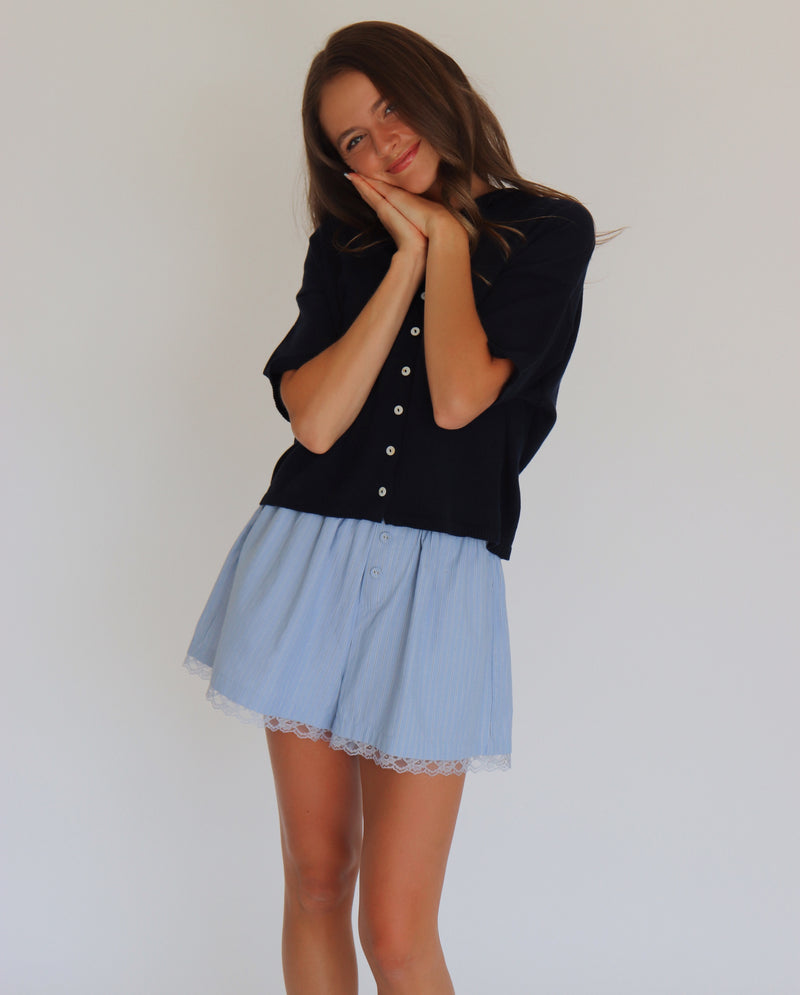 Woman wearing a navy short sleeve cardigan and light blue shorts against a white background