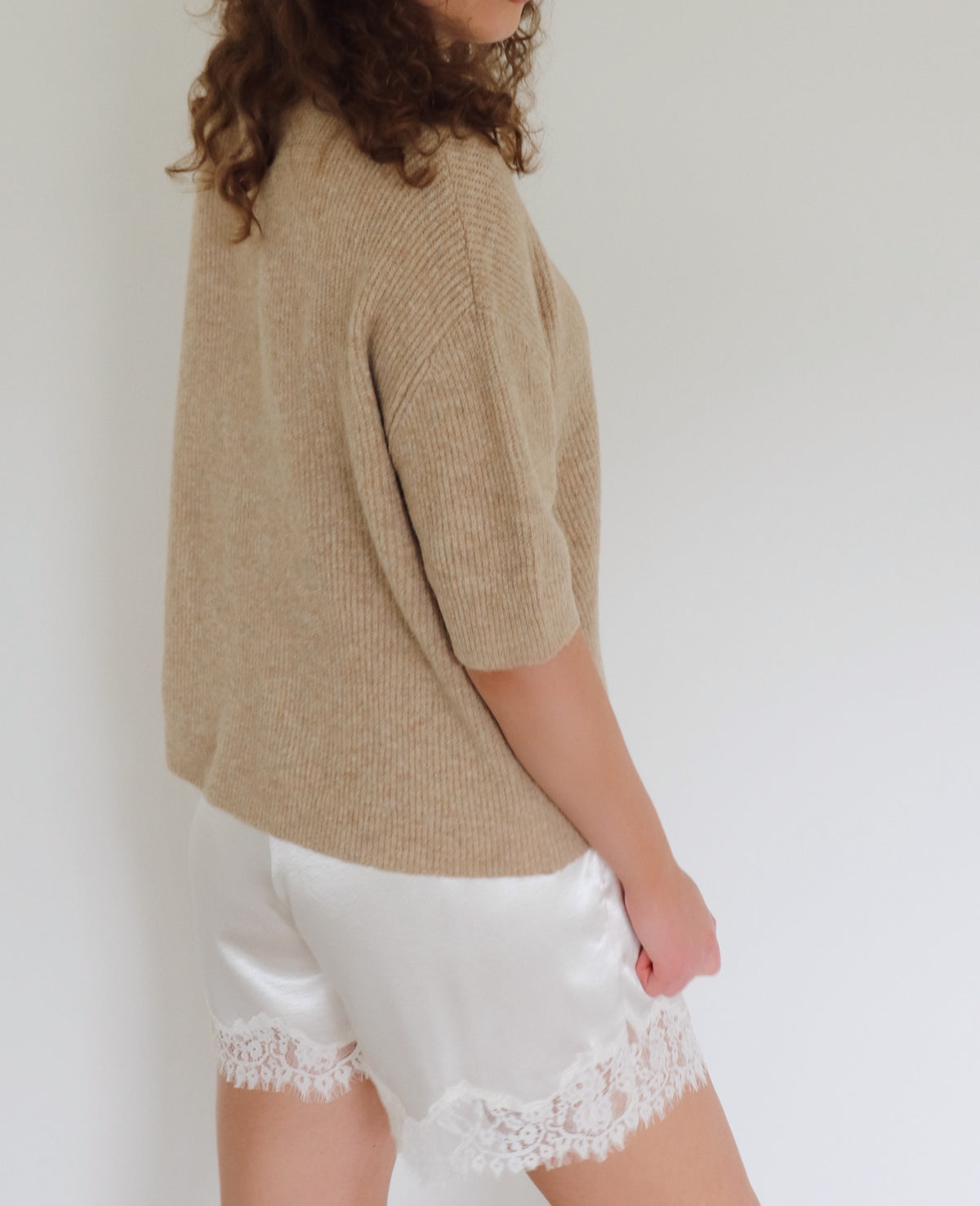 A woman shown from the side wearing a beige knit short-sleeve top and white satin shorts with a lace hem, against a plain light background.