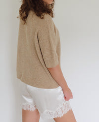 A woman shown from the side wearing a beige knit short-sleeve top and white satin shorts with a lace hem, against a plain light background.