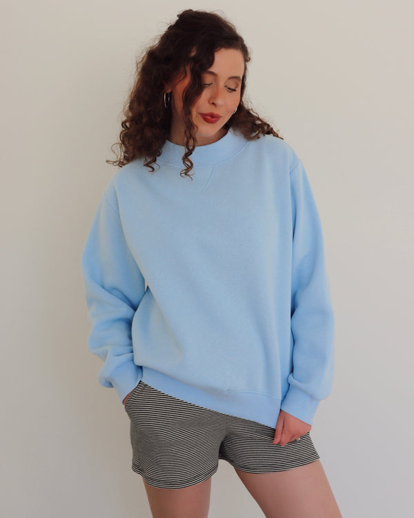 Woman wearing a light blue pullover and striped shorts against a plain background