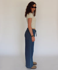 A woman wearing a cream short-sleeve crop top, high-waisted wide-leg blue denim jeans with a drawstring waist, and tan slip-on shoes, standing in profile against a plain white studio background.