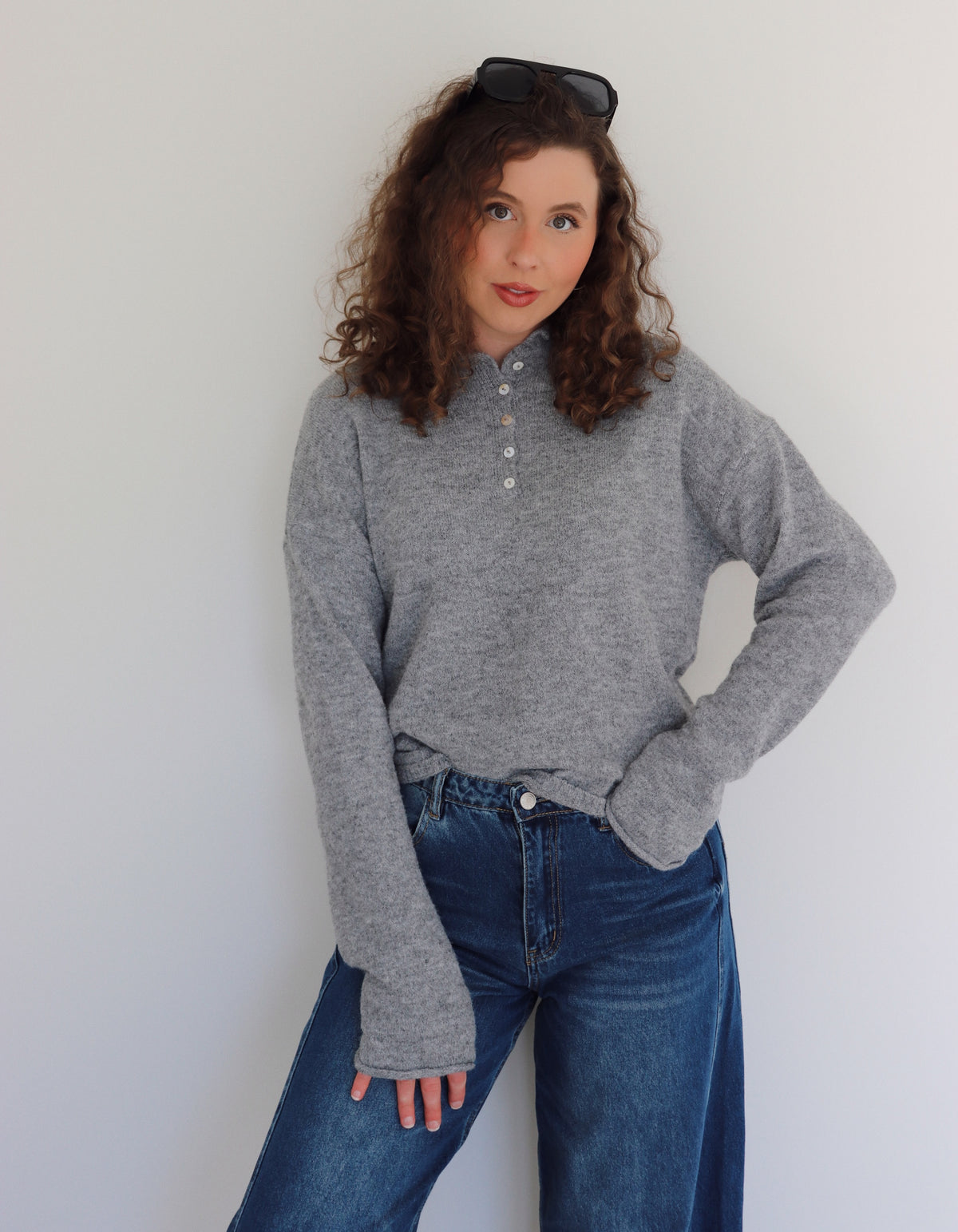 Woman wearing a gray top  and blue jeans against a white wall
