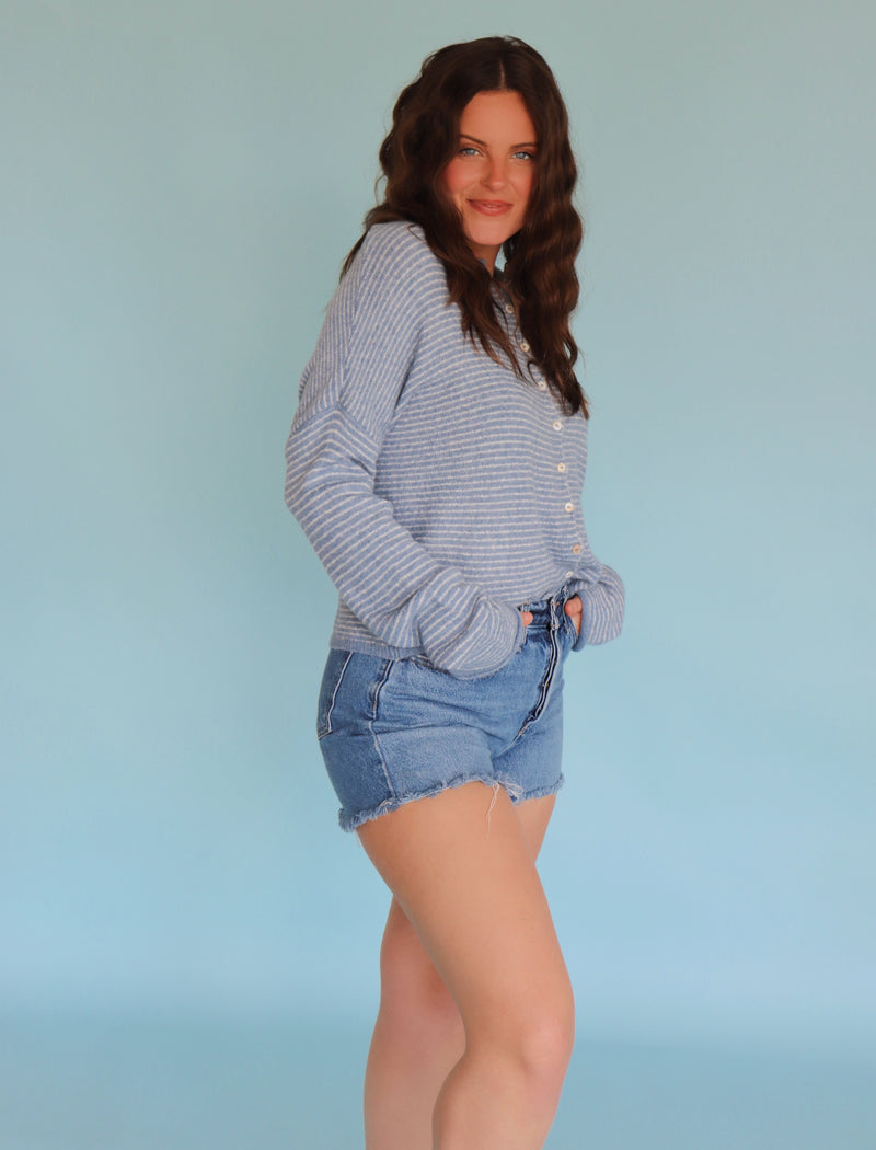 Woman wearing a striped long-sleeve cardigan and denim shorts against a light blue background
