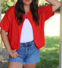 Person wearing a red short-sleeve top over a white shirt with denim shorts outdoors.