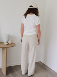 A person wearing loose, wide-leg drawstring pants in a cream, blue and beige stripe pattern, paired with a white top and white sneakers, standing in a minimalist room with wood floors.