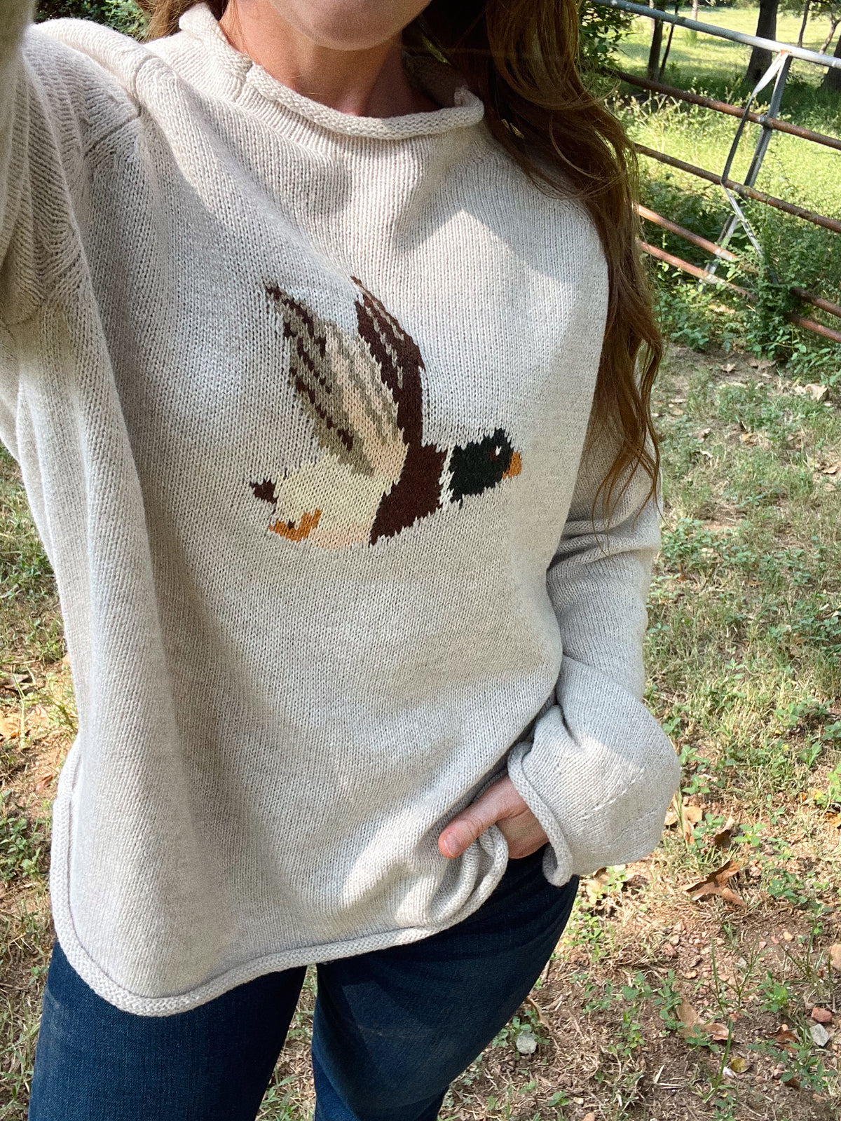 A woman wearing a cream/gray colored knit sweater with a detailed bird motif on the chest, paired with blue jeans, standing outdoors in a wooded setting.
