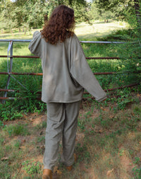 A woman wearing a matching olive green striped lounge set stands outdoors in a wooded area with greenery and a rustic fence behind her.