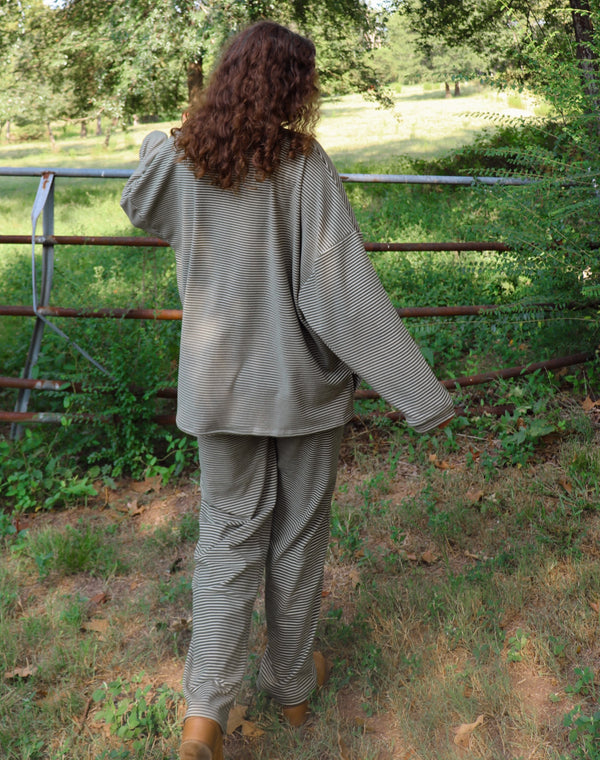 A woman wearing a matching olive green striped lounge set stands outdoors in a wooded area with greenery and a rustic fence behind her.