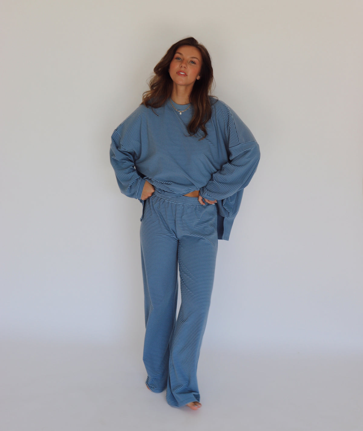 A woman modeling a matching blue striped lounge set, consisting of a long-sleeve top and wide-leg pants, against a plain white studio background.