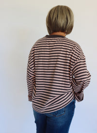 A woman with a short blonde bob wearing a brown and gray striped long-sleeve top and blue jeans, standing against a plain white wall.