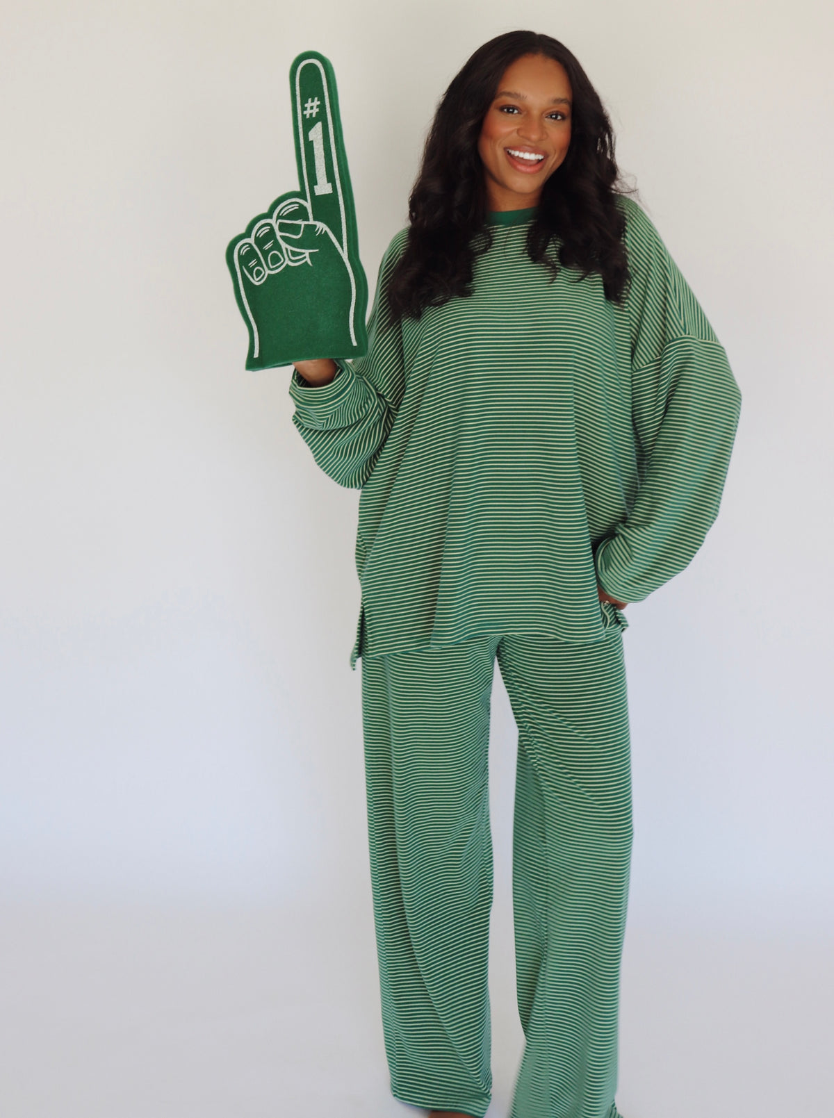 A woman modeling a matching green and yellow striped lounge set, featuring a long-sleeve top and wide-leg pants, while holding a green foam finger with white text.