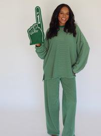 A woman modeling a matching green and yellow striped lounge set, featuring a long-sleeve top and wide-leg pants, while holding a green foam finger with white text.