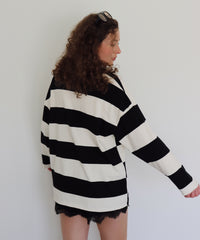 A woman with curly brown hair wearing an oversized black and off-white striped long-sleeve sweater and black lace-trim shorts, standing against a plain light gray wall.